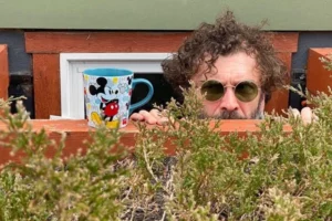 a caucasian man with medium length curly brown hair wearing round wire rim sunglasses peers over the top of a wall with a Mickey Mouse coffee mug sitting atop the ledge and bushes in the foreground