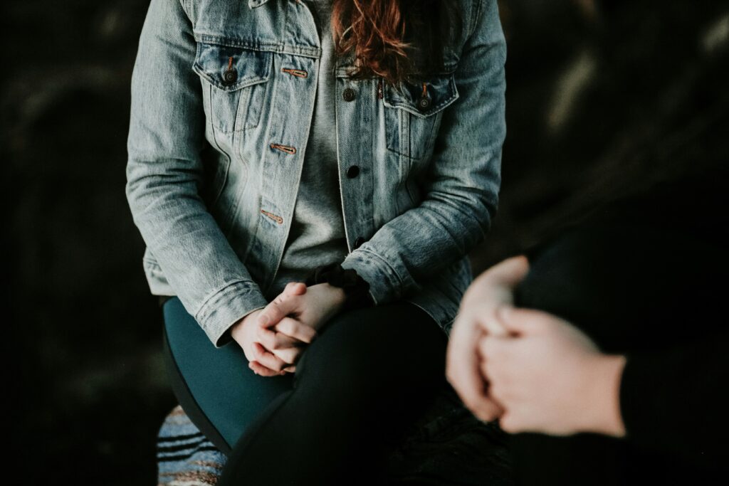 a white woman with red hair in a jean jacket sits with her hands clasped and legs crossed, talking to another person with clasped hands