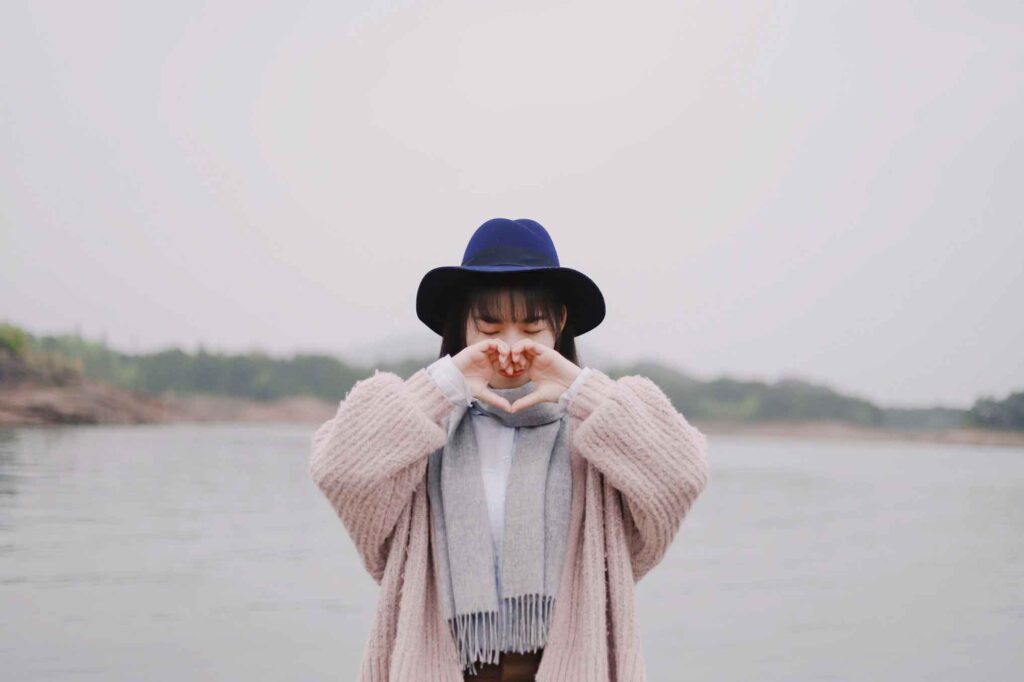 a young asian woman with bangs in a blue boho hat and pink cardigan makes a heart shape with her hands while standing in front of a lake