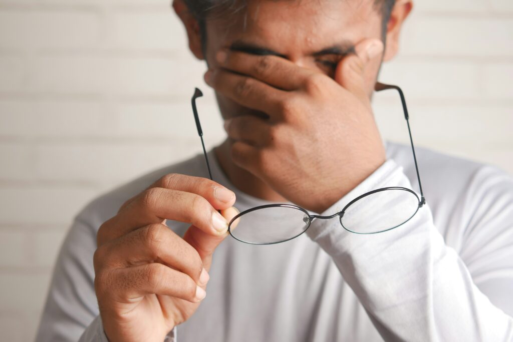 a young man holds his glasses in front of his chest while he rubs his eyes with the other hand