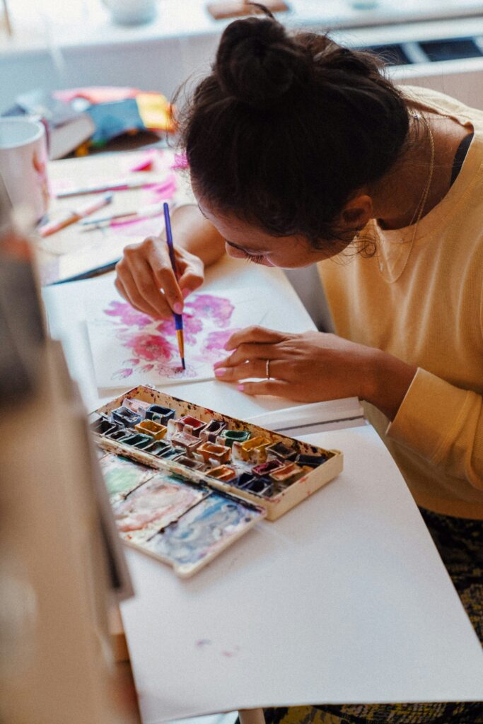 overhead view of a woman with her dark hair in a bun and yellow sweatshirt painting flowers on white paper