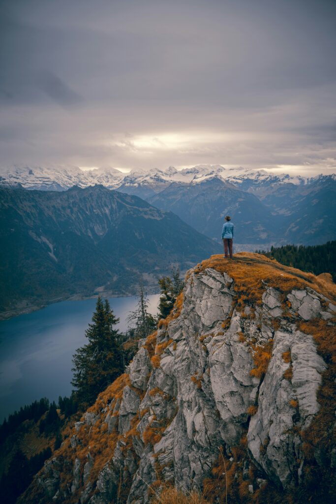 a person in a blue shirt and dark pants stands on top of a cliff in the distance overlooking a river below with snow capped mountains in the background